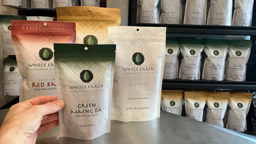 A hand setting a pouch of Whole Earth Green Maeng Da kratom capsules on the counter at Whole Earth Gifts in Arlington, Texas, with stocked shelves behind.